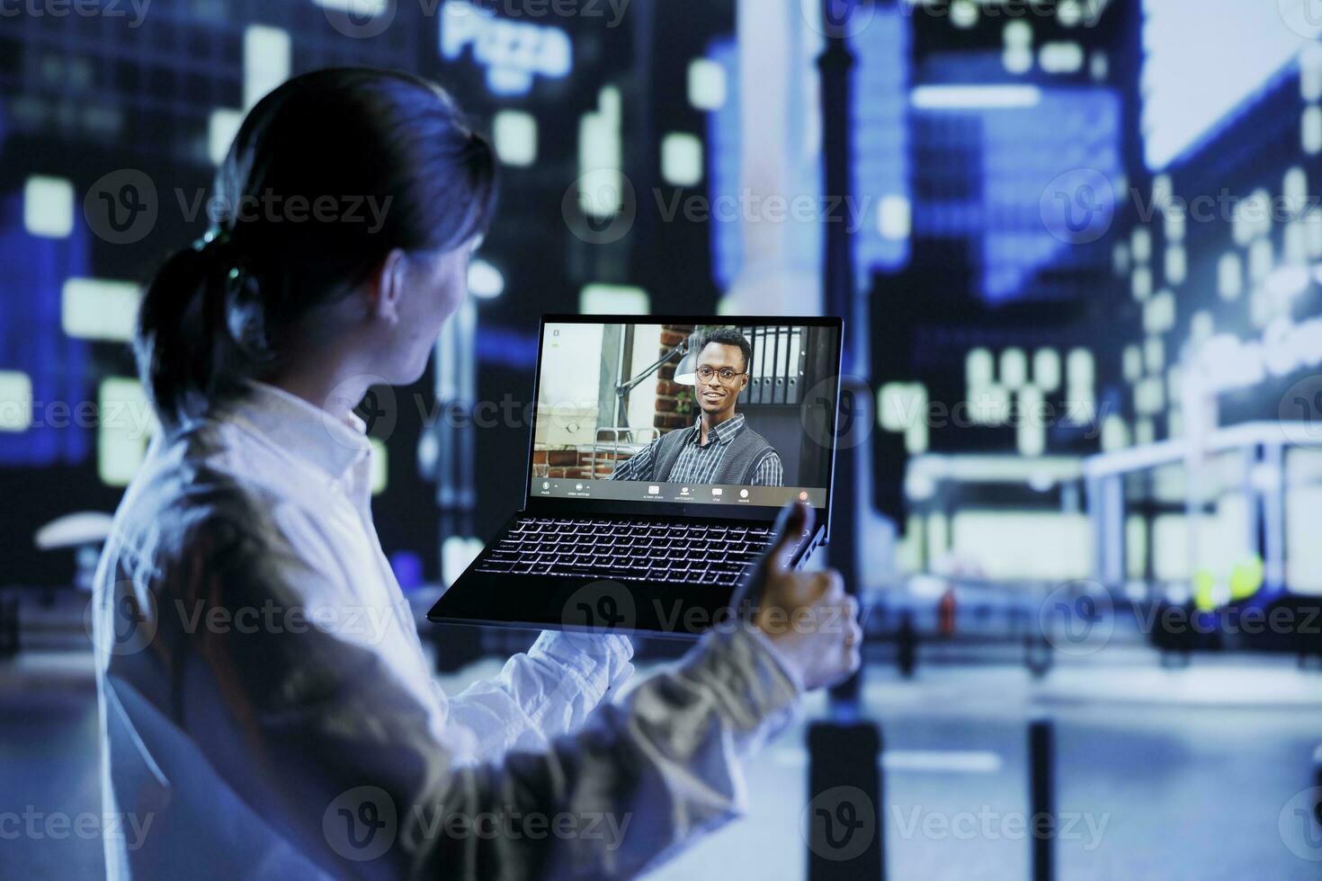 Asian man in videocall with coworker using device while strolling around city streets at night, giving him work updates. Person uses laptop to show colleague surroundings in illuminated urban district photo