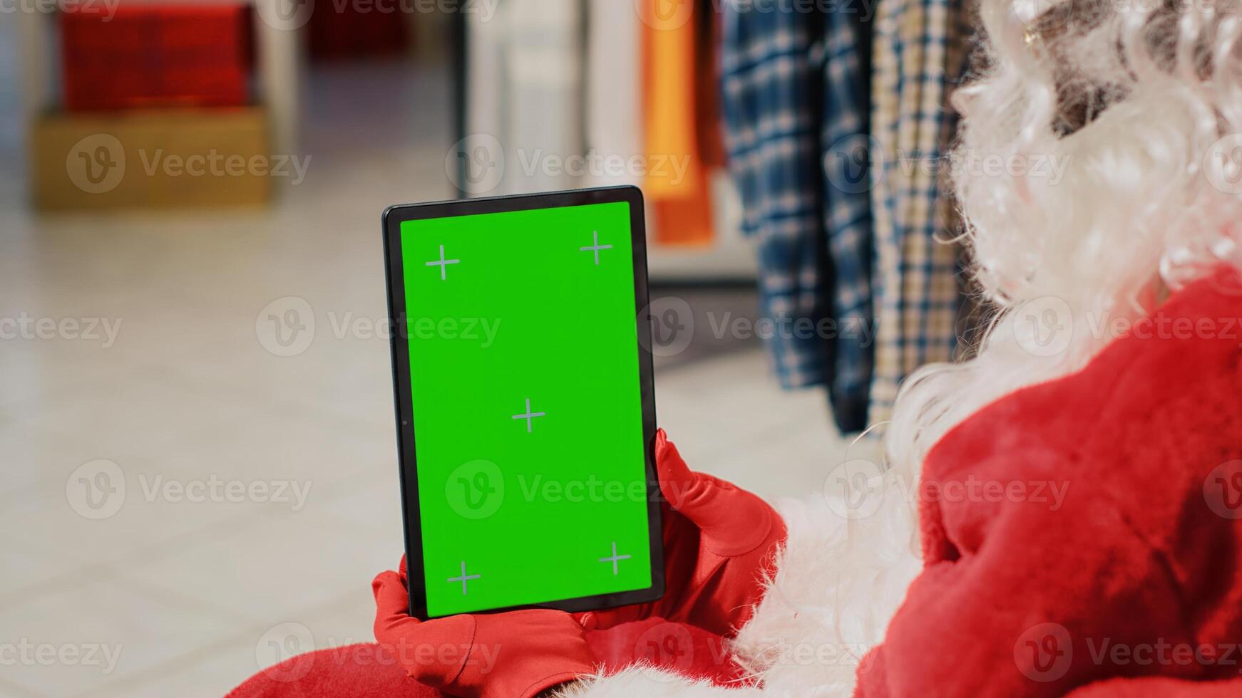 Employee in festive decorated clothing store dressed as Santa Claus holding chroma key tablet, setting up website clothing articles, inputting promotional xmas sales online photo