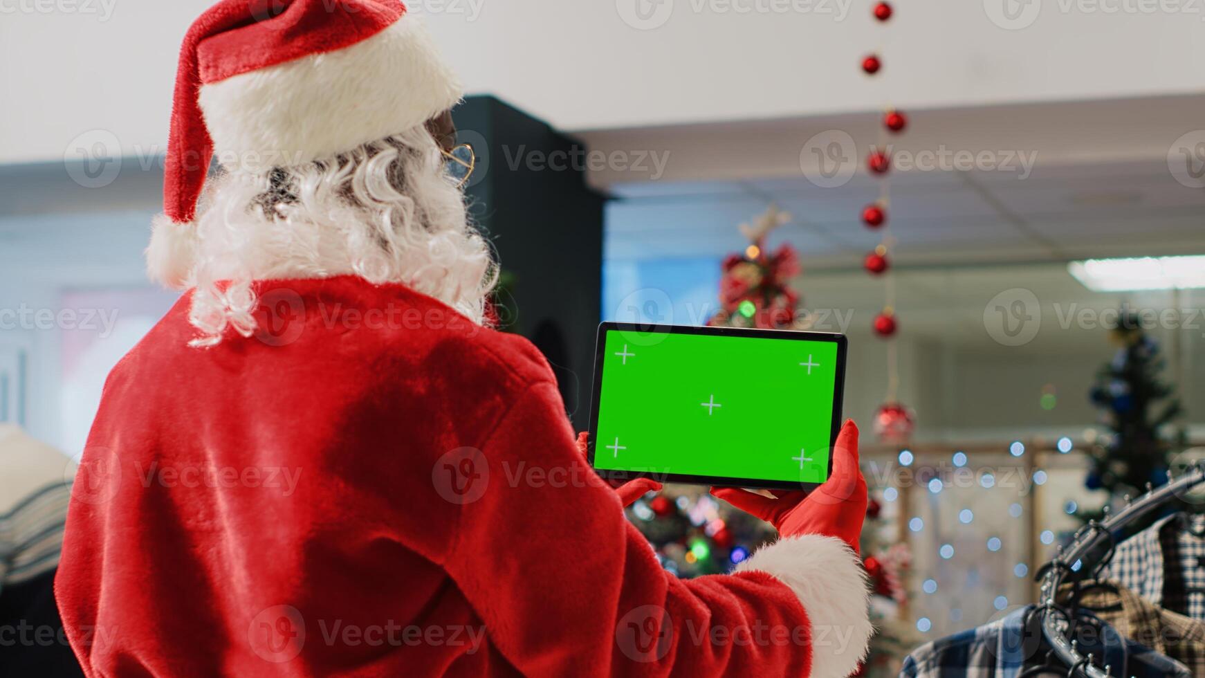 Christmas ornate shopping mall employee impersonating Santa Claus holding mockup tablet, taking a break from job to watch videos on chroma key screen while clients walk around, close up photo