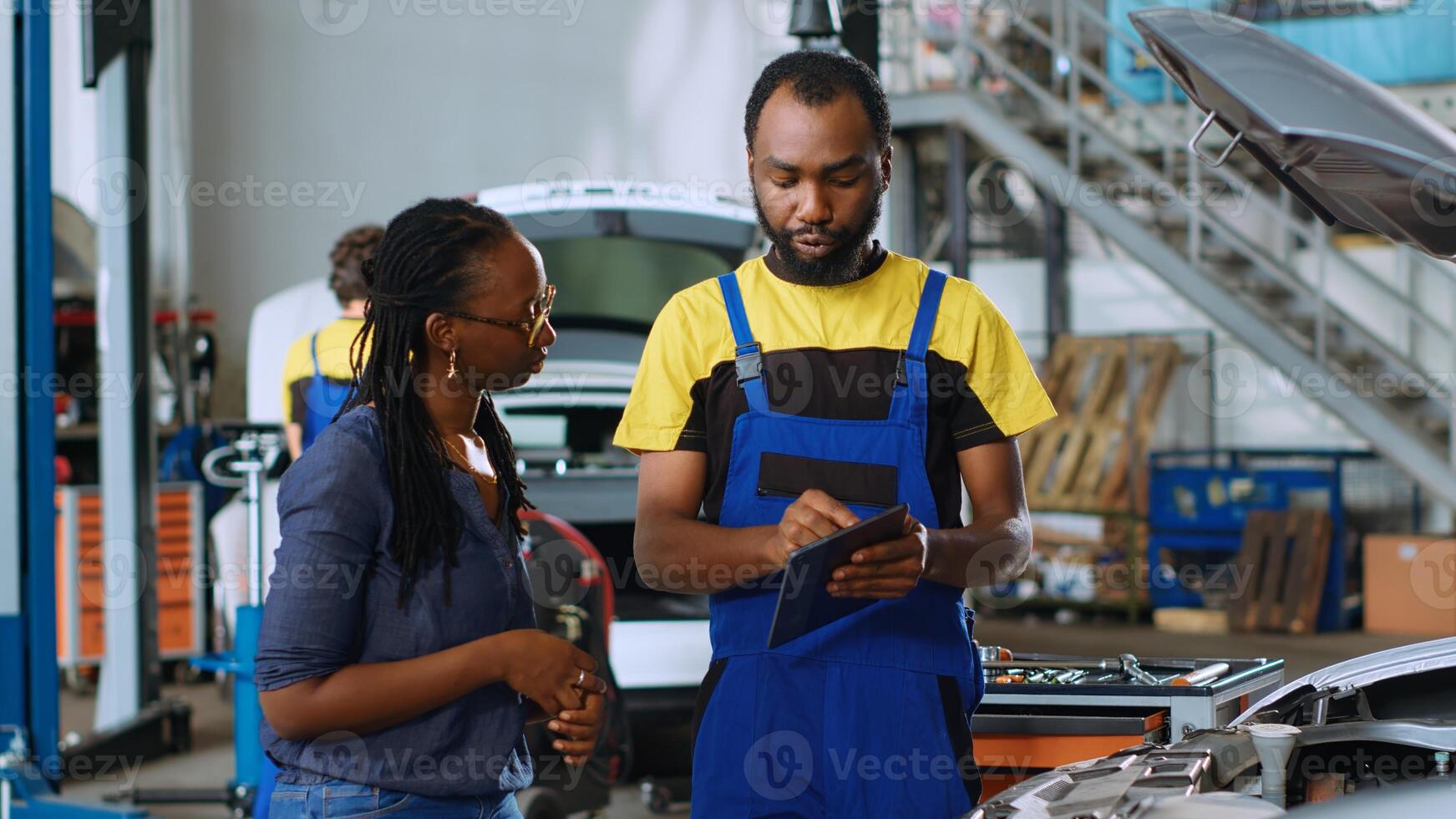 Seasoned auto repair shop mechanic showing client defects found inside car after checkup. African american professional telling woman about unfunctional battery found in her vehicle photo