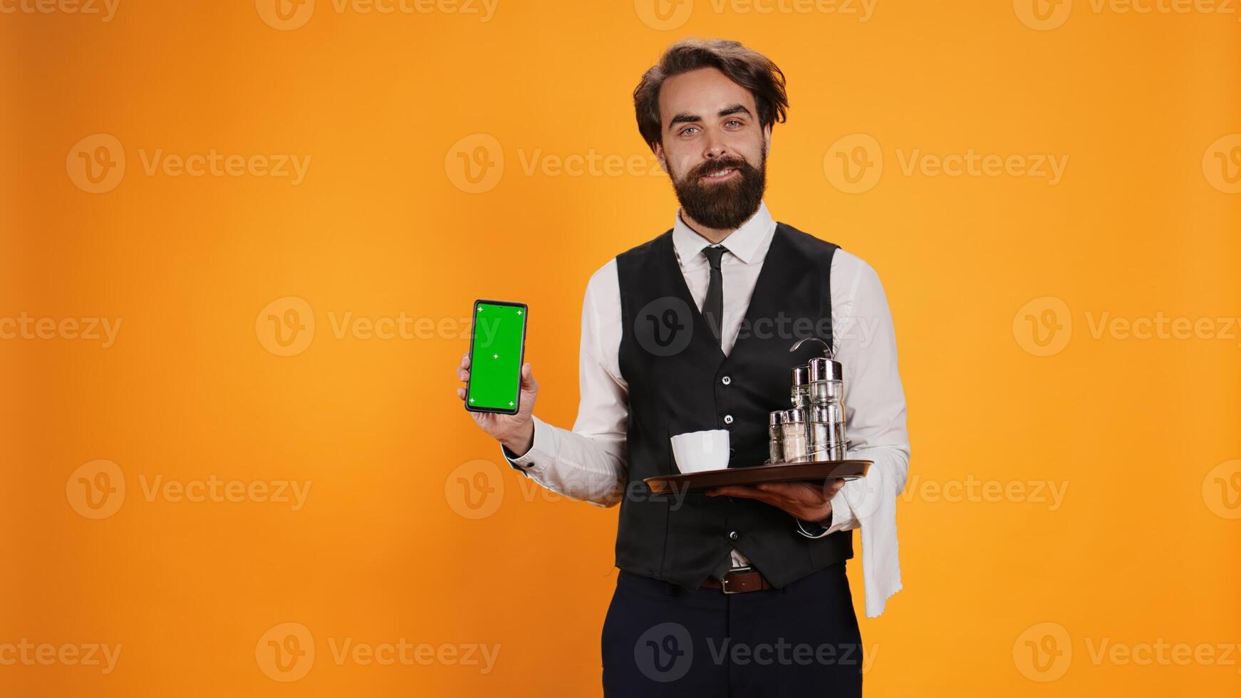 Bearded waiter presents greenscreen on phone display, holding smartphone to show isolated mockup screen. Elegant skilled restaurant valet using empty copyspace template in studio. photo