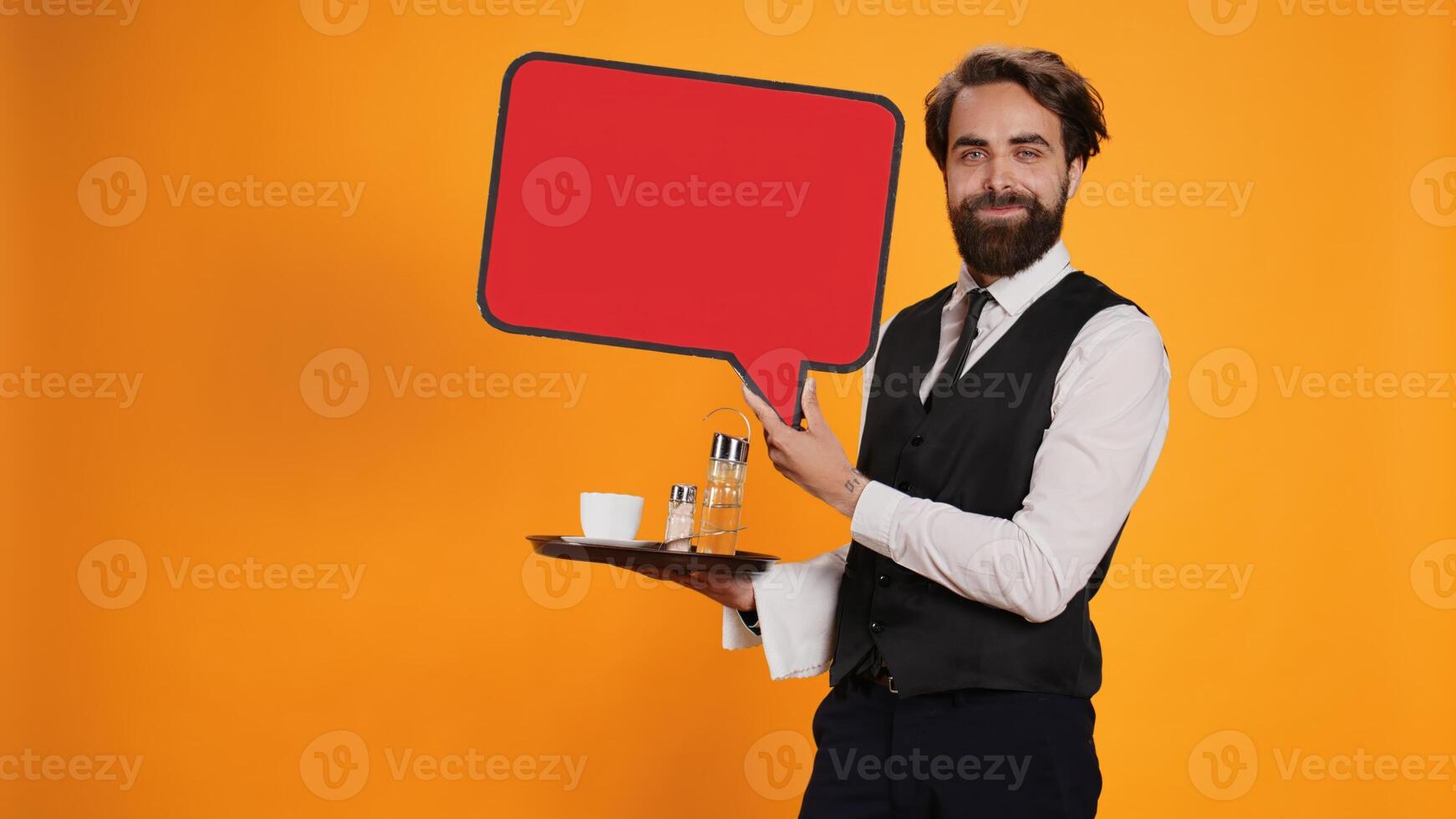 Elegant waiter holds speech bubble icon on camera, working in restaurant industry to create marketing ad with red cardboard cutout. Employee presenting billboard with blank isolated space. photo