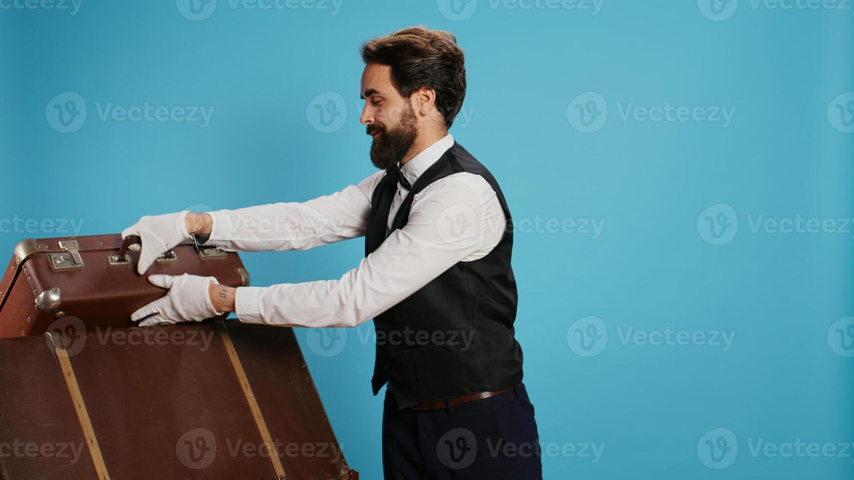 Classy hotel employee with baggage posing against blue background to advertise tourism or travel field. Bellboy working in hospitality with formal clothing attire, carrying trolley bags. photo