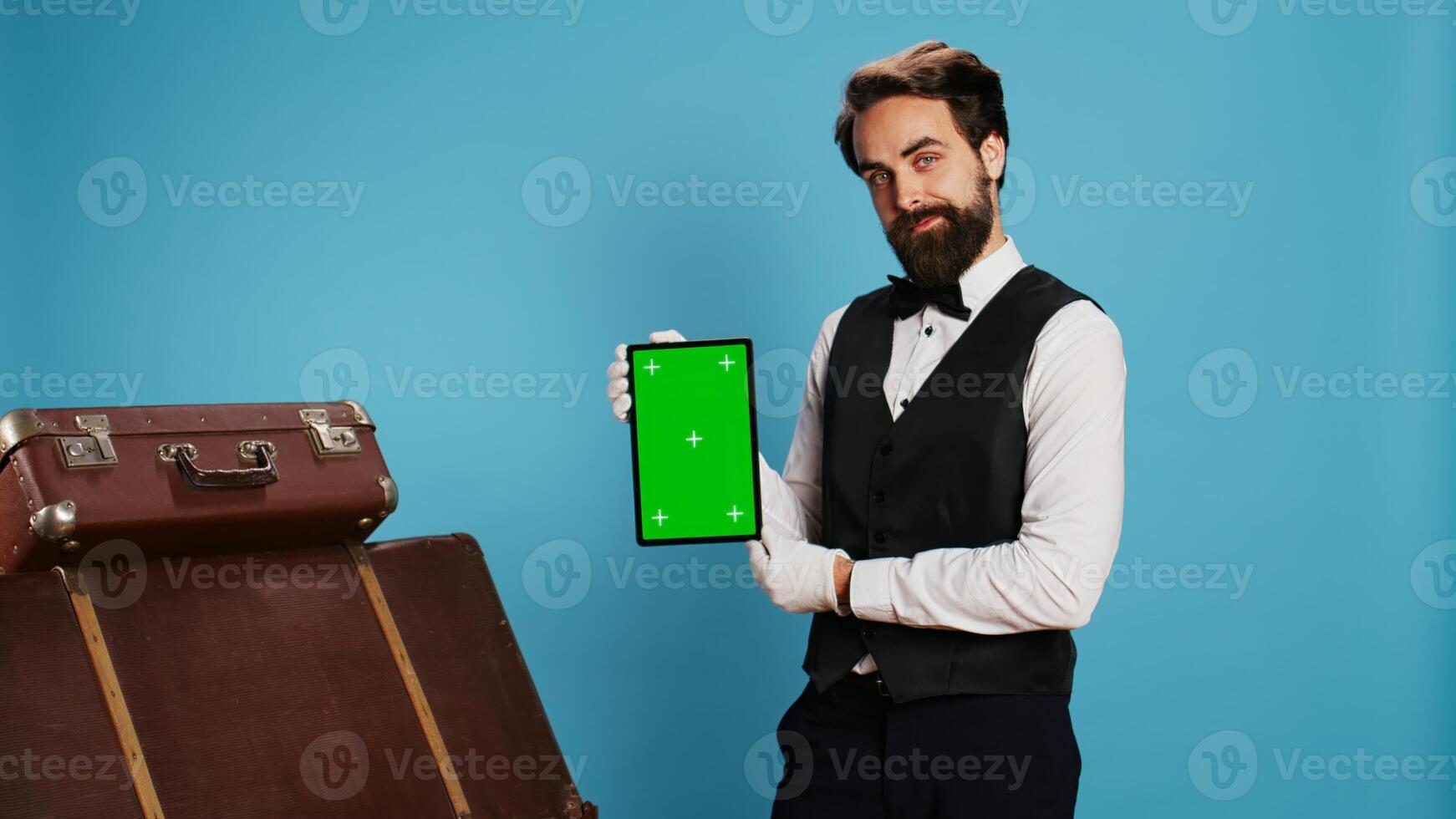 Doorman poses with greenscreen tablet on camera, standing next to blue background in studio. Hotel worker showcasing isolated mockup copyspace on device, hospitality concept. photo