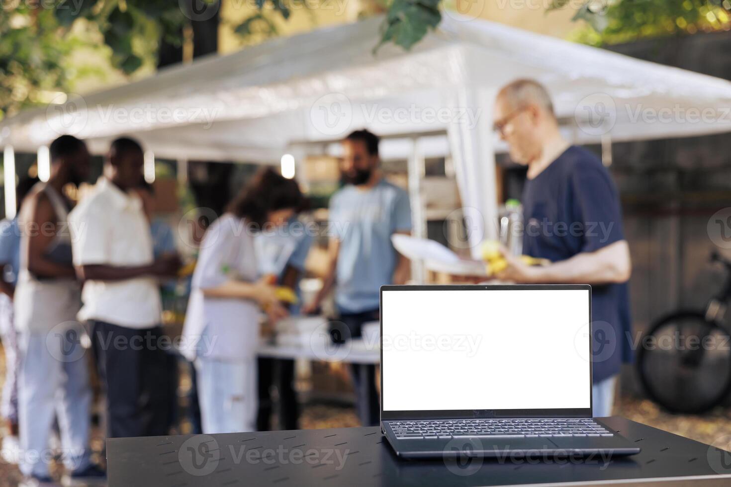 Picture shows digital laptop with a blank white screen that is sitting on a table while charity workers help the less fortunate. Minicomputer in the foreground with isolated copyspace mockup template. photo