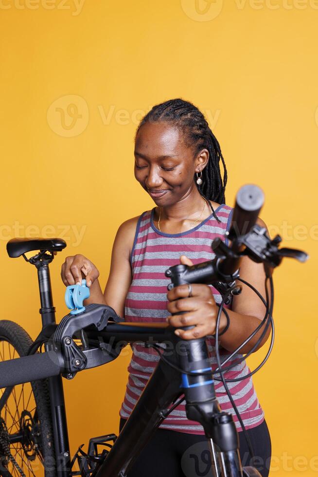 Black female cyclist inspecting her bicycle broken frame while placed on repair stand against isolated backdrop. Young woman preparing to use specialized tools for adjustments and repairs. photo