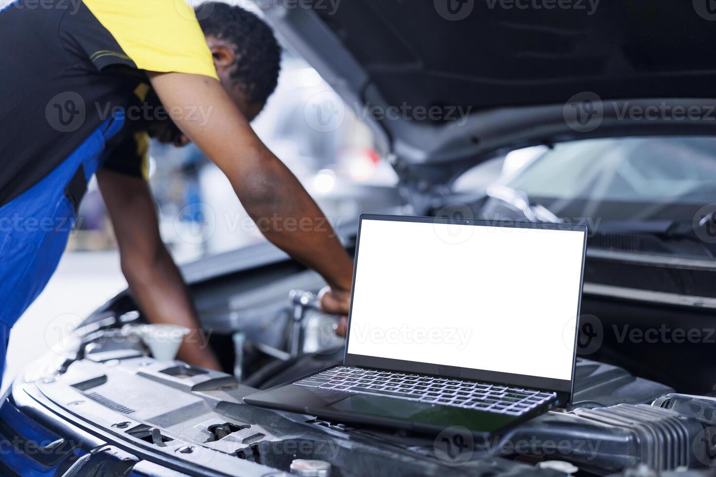 Mockup laptop on car with hood open while BIPOC repairman in blurry background replaces brakes. Isolated screen device next to experienced garage worker fixing customer automobile photo