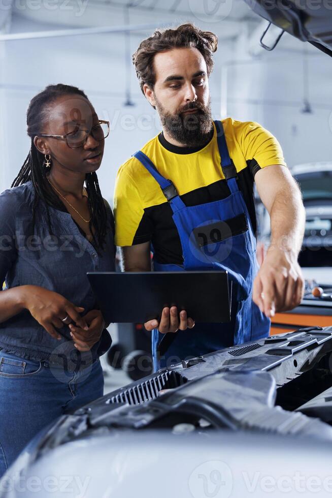 Mechanic in car service uses laptop to calculate invoice after fixing broken automobile distributor. Certified auto repair shop worker uses device to inform woman of final costs after mending vehicle photo