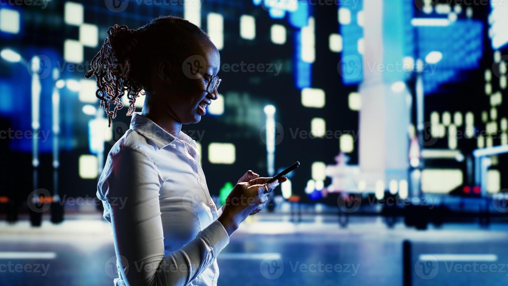 Joyful woman discussing with her friends using smartphone while wandering around dimly illuminated city streets at night. Chipper girl walking outside in urban environment, texting people photo