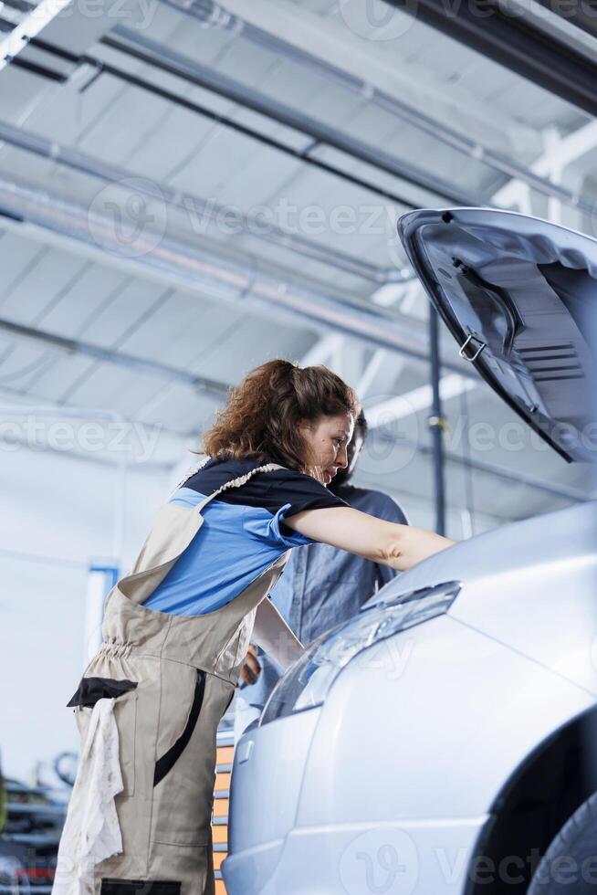 Engineer in garage finishing fixing car for african american customer, looking underneath vehicle hood to remove remaining oil leaks. Worker does routine engine cleaning on client automobile photo
