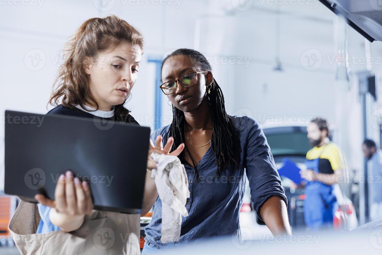 Mechanic in car service uses laptop to calculate invoice after fixing broken automobile radiator. Skilled garage worker uses device to inform woman of final costs after mending vehicle photo