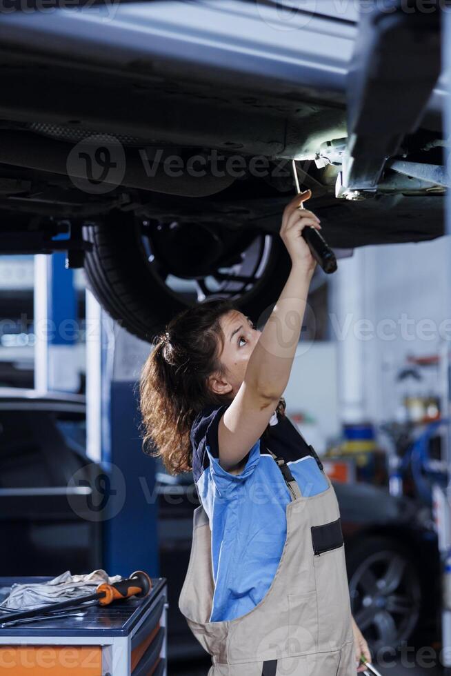 Seasoned mechanic working on suspended car in garage, checking engine during routine maintenance. Auto repair shop professional underneath vehicle, inspecting components using work light photo