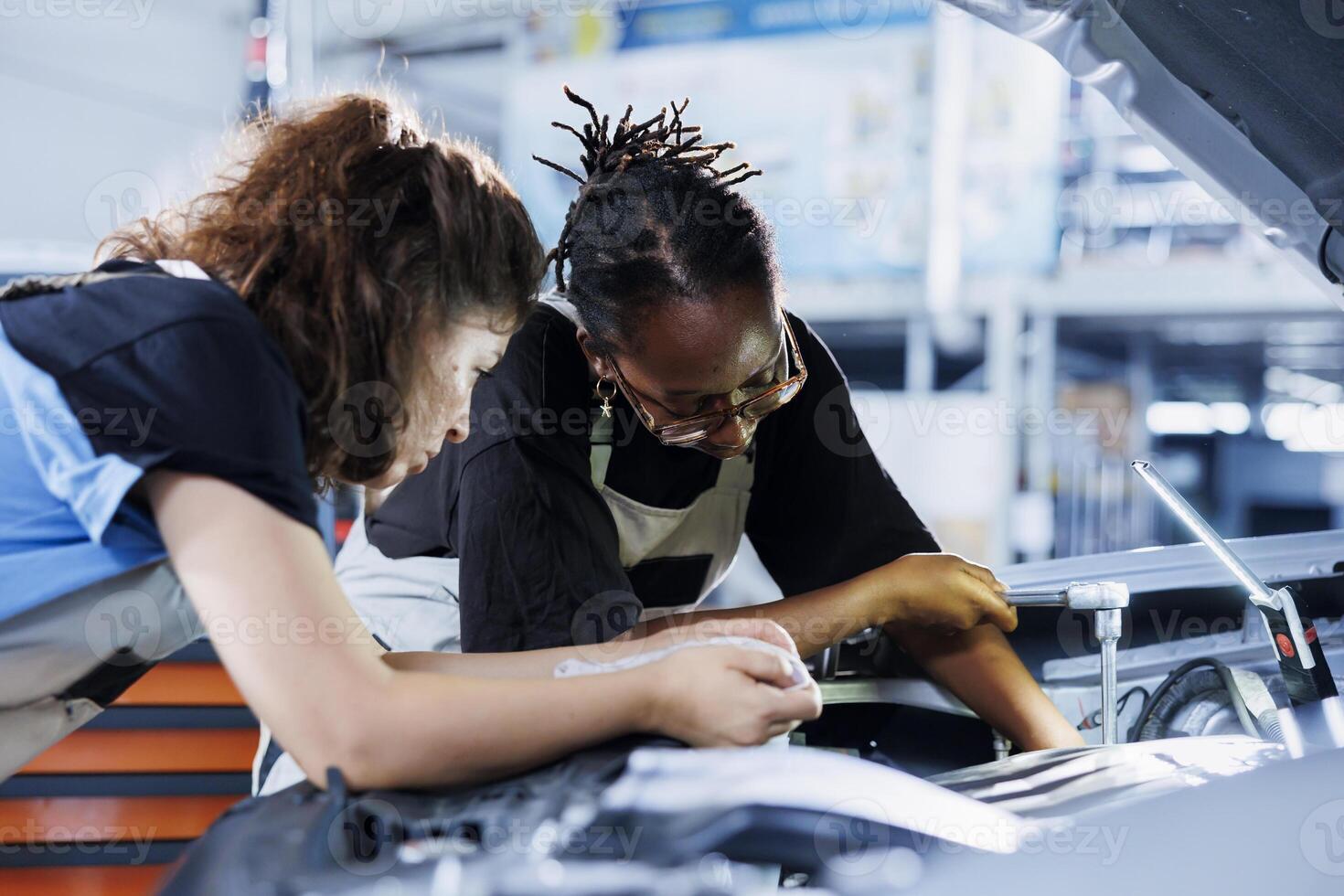 Team of women in auto repair shop working together on fixing car, discussing best options. Teamworking mechanics collaborating on servicing broken vehicle, checking for damaged fuel tank photo
