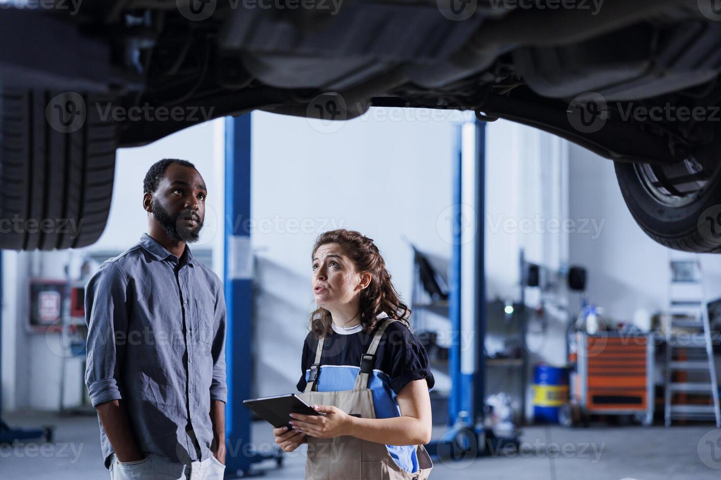 Mechanic at auto repair shop conducts annual vehicle checkup, informing client about needed tires replacement. Garage worker talks with customer underneath suspended car after finishing inspection photo