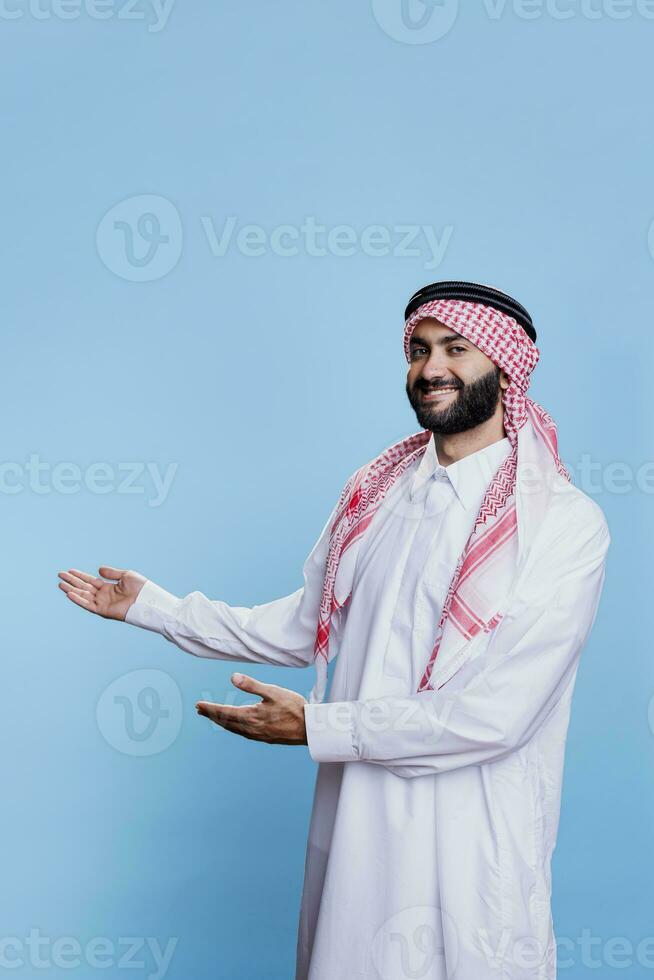 Excited arab sales person dressed in traditional islamic clothes advertising product, pointing to side with hands. Cheerful muslim man showing copy space and looking at camera photo
