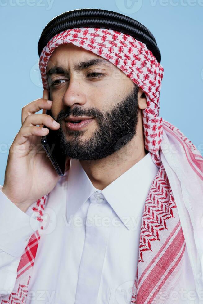 árabe vistiendo ghutra tocado con igual cuerda banda y chateando en teléfono inteligente de cerca. musulmán hombre vestido en tradicional ropa Hablando en móvil teléfono en estudio de cerca foto