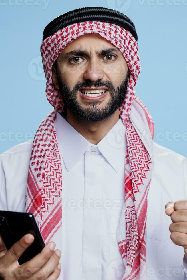 Excited man dressed in traditional muslim clothes saying yes while using smartphone and looking at camera with clenched fist. Arab in thobe holding mobile phone and posing in studio photo