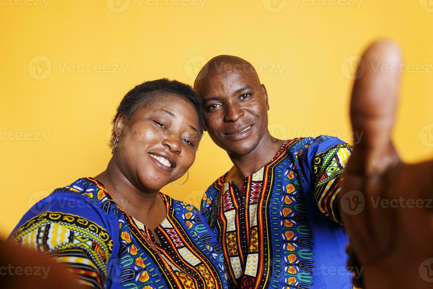 Smiling black man and woman wearing ethnic clothes taking selfie on smartphone and looking at front camera. Cheerful couple holding mobile phone and making photo pov portrait