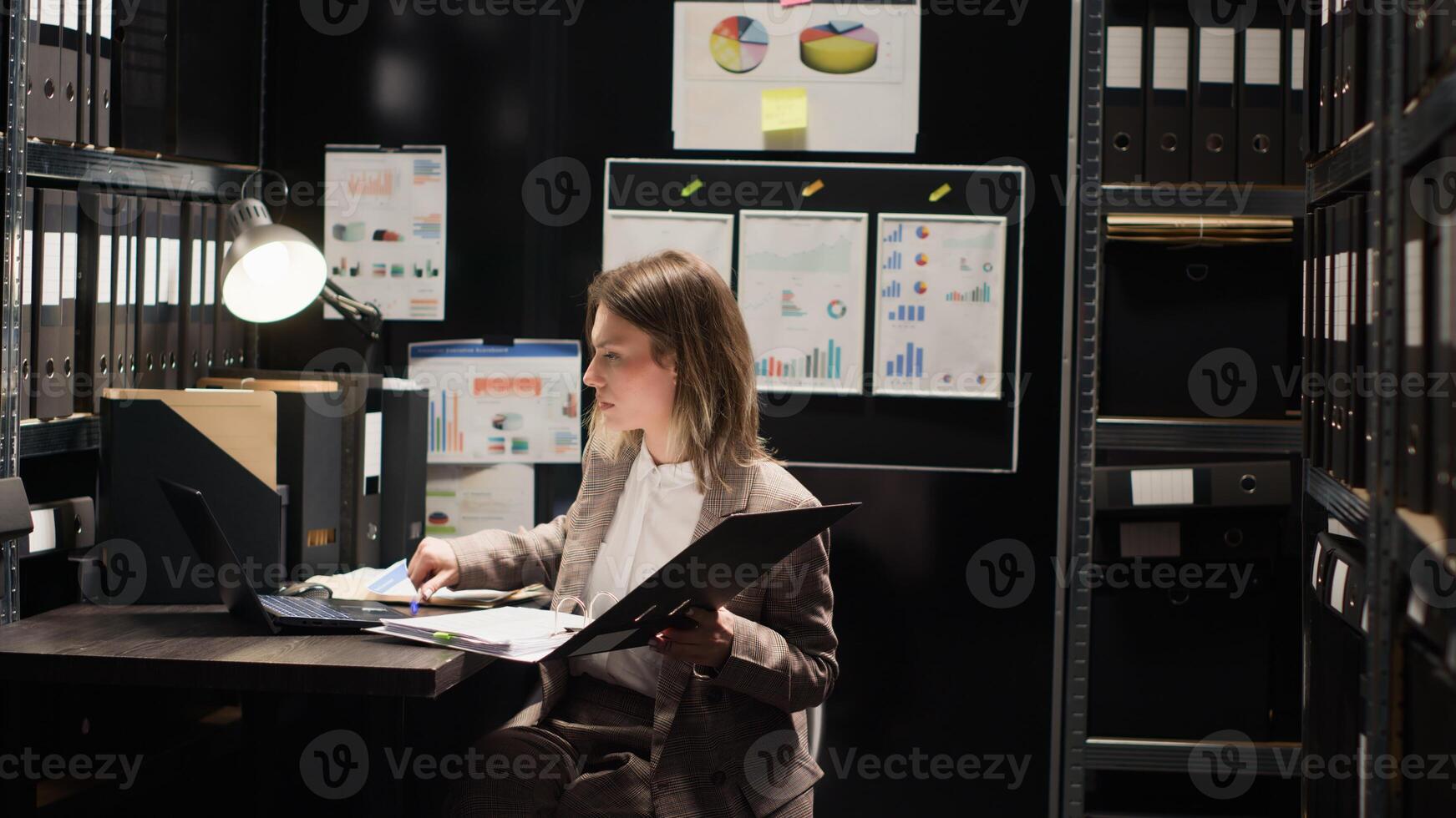 Private investigator enters office with case folder, examining evidence on desk with files and laptop. Caucasian woman with expertise in criminal investigation, reviews statements to identify suspect. photo