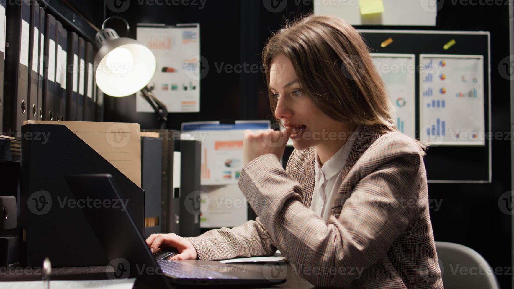 Focused female investigator conducting investigation and reviewing evidence. Zoom-in shot of inspector sitting at desk with laptop and case files for comparing statements and profile of the suspect. photo