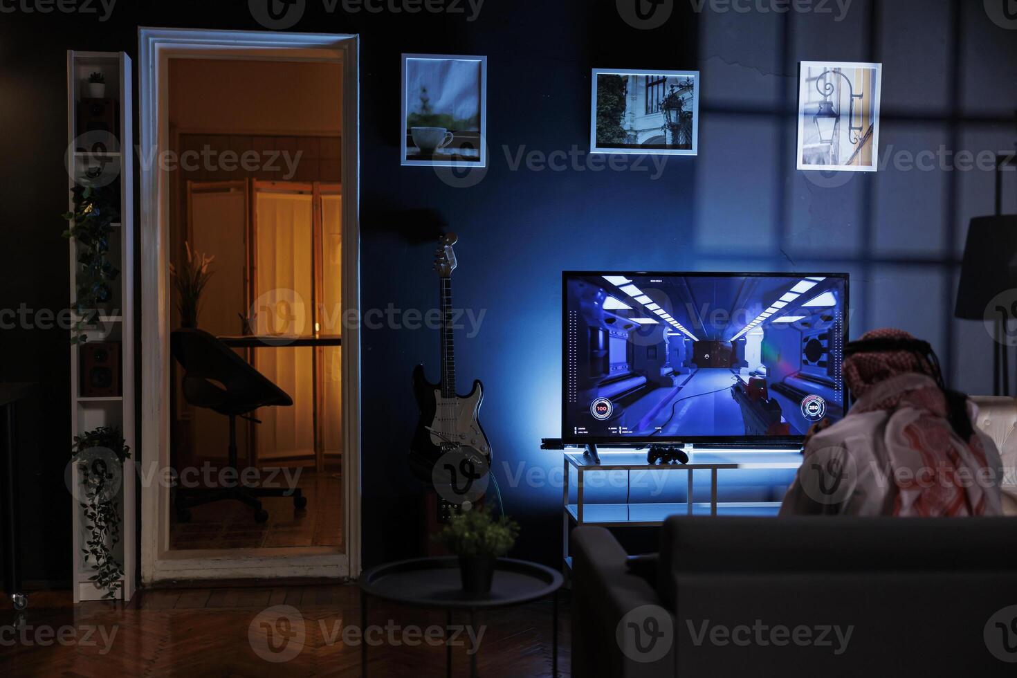 Rear-view shot of Muslim man seated on the sofa engrossed in playing video games. Image shows a Middle Eastern man dressed in traditional clothing enjoying online entertainment from the television. photo