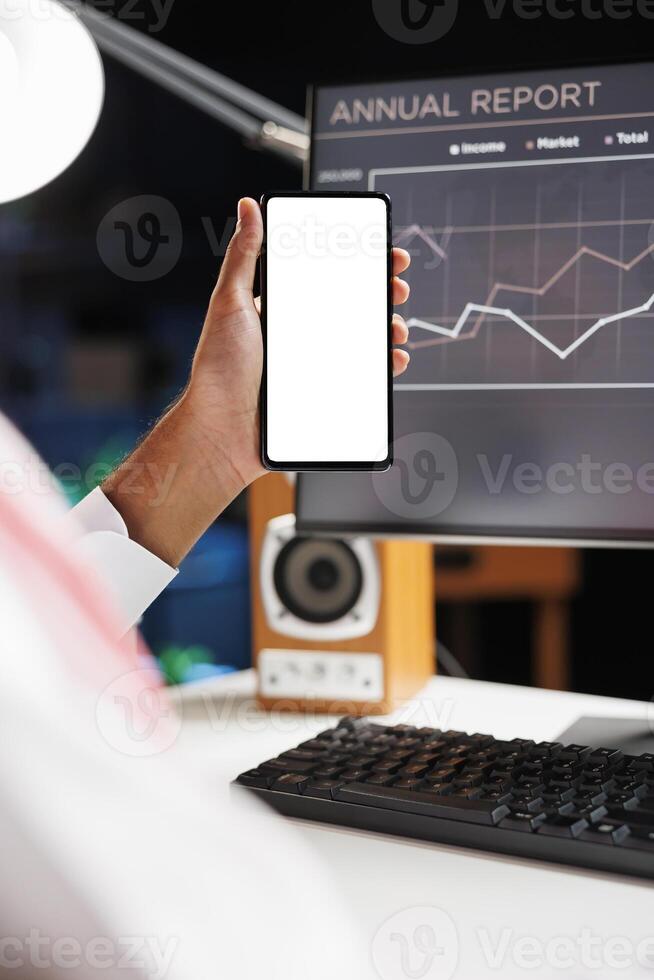 Selective focus of a guy hand holding a cellphone in front of a desktop computer with an isolated copyspace white screen. Arab showing a mobile device with a blank chromakey mockup template. photo