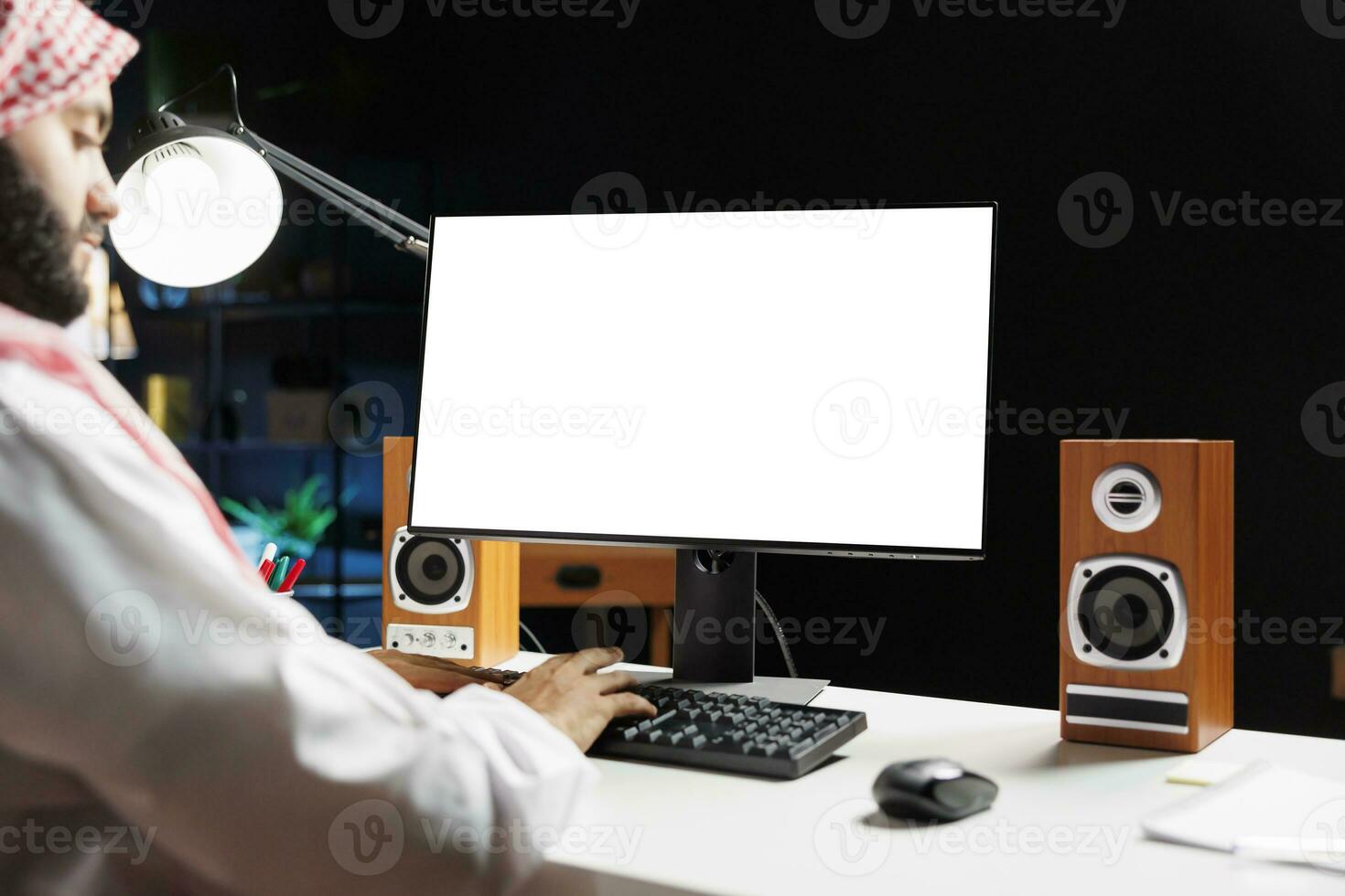 imagen retrata musulmán chico sentado a su puesto de trabajo a hogar, su computadora monitor demostración un aislado chromakey modelo. juvenil árabe hombre trabajando con un escritorio ordenador personal mostrando copyspace blanco pantalla. foto