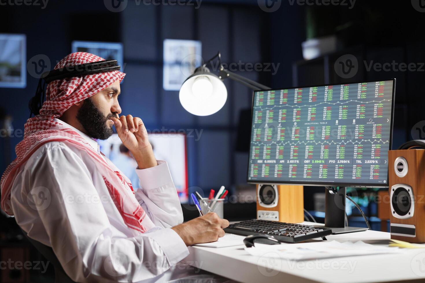 Diligent Muslim man working at a desk with a computer, checking financial reports and taking notes. Focused Arab guy using desktop pc, working with documents at the home office. photo