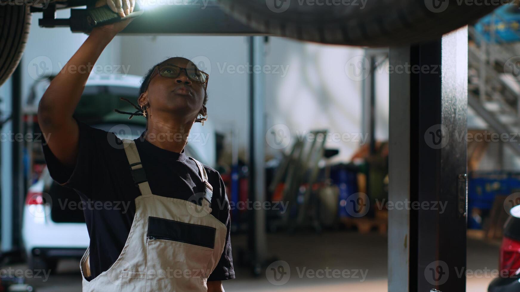 Trained engineer underneath vehicle on overhead lift in garage, checking parts . African american car service professional using work light to make sure automotive underbody is in proper condition photo