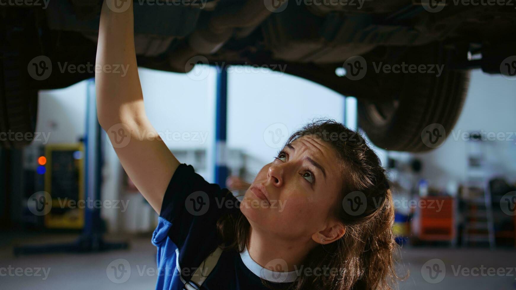 Qualified engineer working on suspended car in garage, checking components during routine maintenance. Auto repair shop employee underneath vehicle using work light to look for potential issues photo