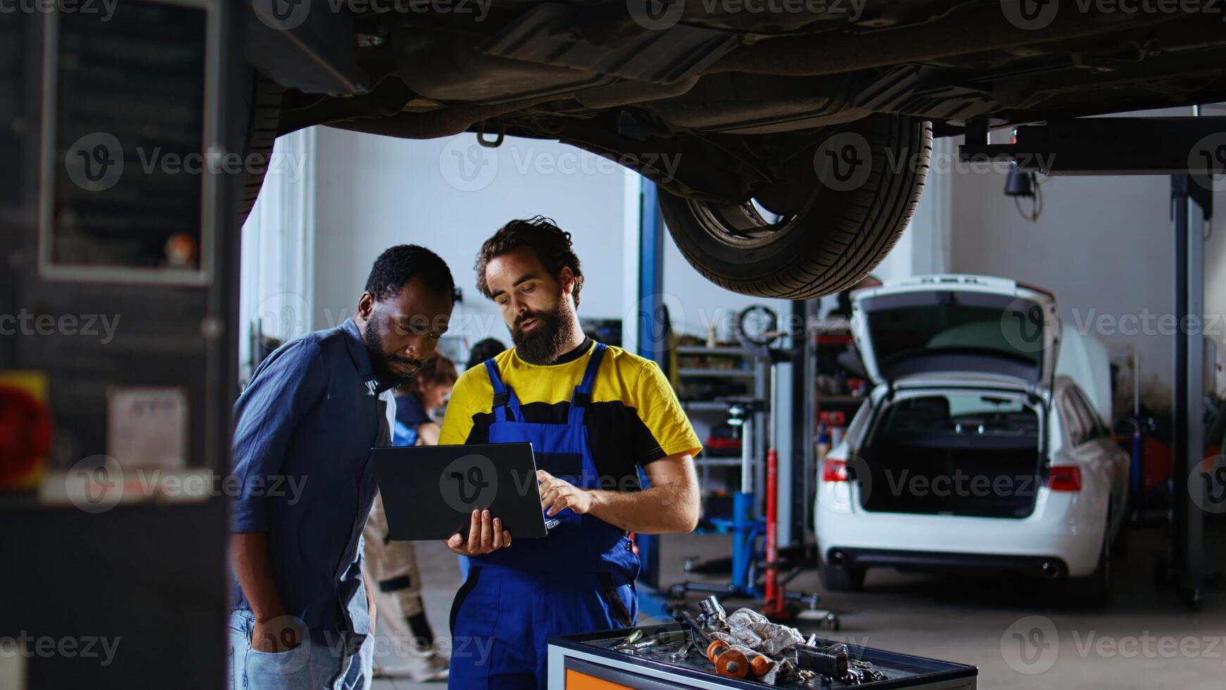 Trained mechanic and customer standing underneath car in garage using laptop to order new parts after finding defects. Employee assisting african american client by looking for components online photo