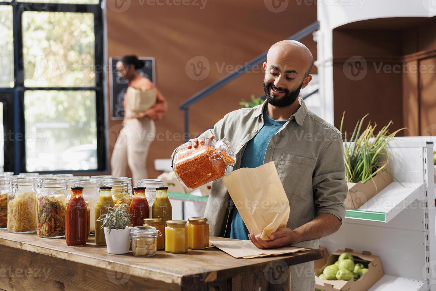 Smiling man pouring orange lentils into bag, buying healthy goods from reusable glass jars with organic bulk products. Customer purchasing natural bio food items from local eco zero waste store. photo