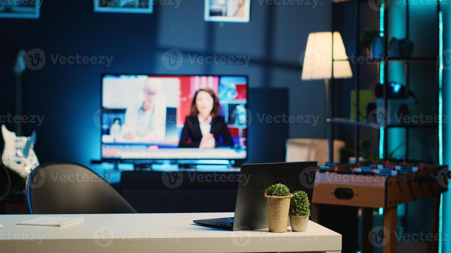 Panning shot of work desk with laptop and mini house plants in dimly lit empty home studio interior. Apartment illuminated by RGB lights with TV news broadcast running in background photo