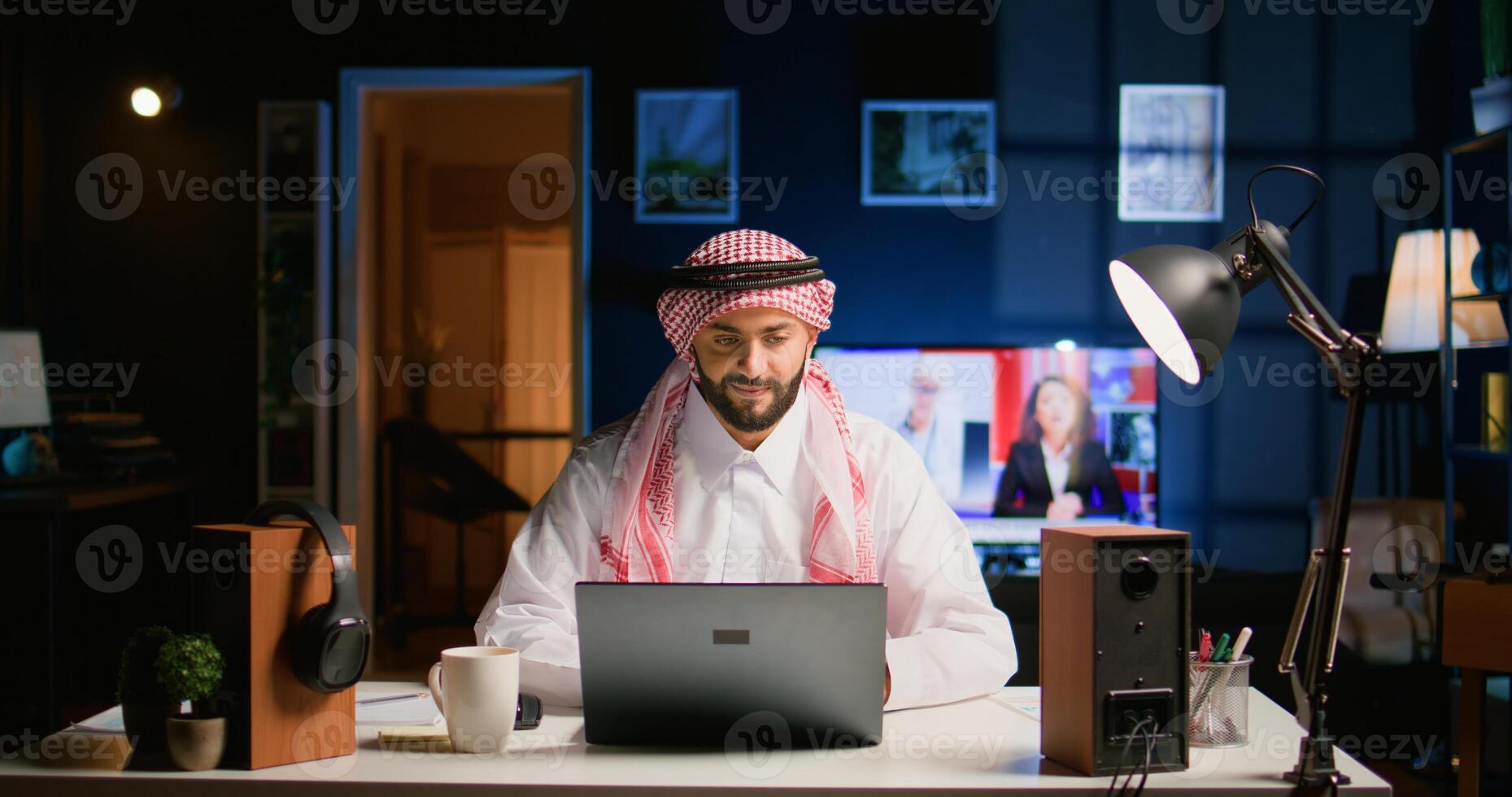 Muslim teleworker remotely working in dimly lit apartment, enjoying fresh cup of coffee. Middle Eastern worker at home typing on notebook device, sending emails and drinking hot beverage photo