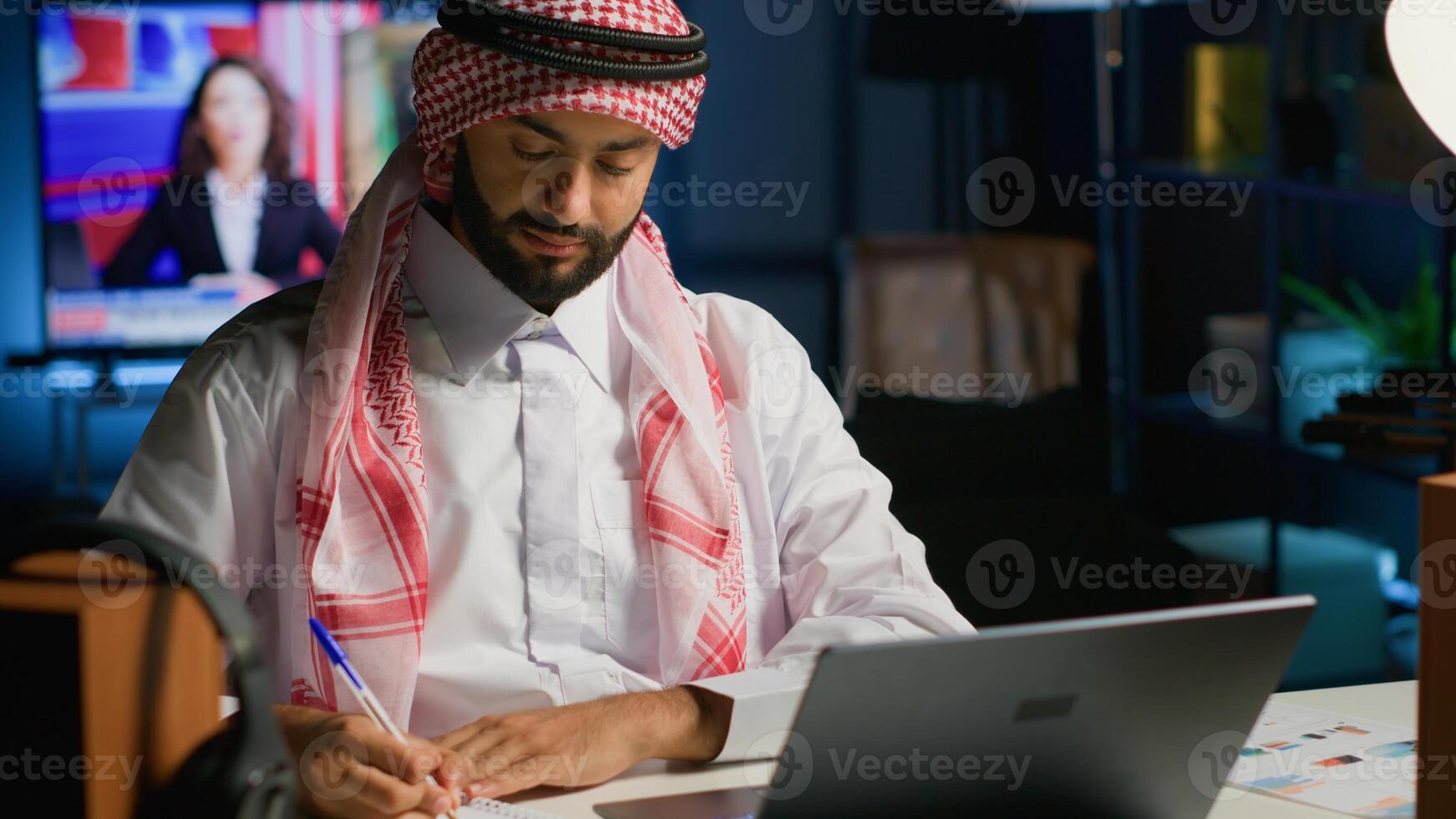 Arab entrepreneur working at home office desk on laptop, browsing the internet and writing in notebook. Muslim man using pen, notepad and digital device while teleworking photo