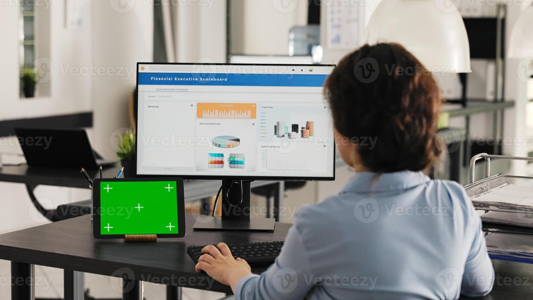 Businesswoman works at desk with greenscreen presented on modern tablet, looking at isolated copyspace template while she analyzes revenue data on pc. Worker uses chromakey display. Handheld shot. photo