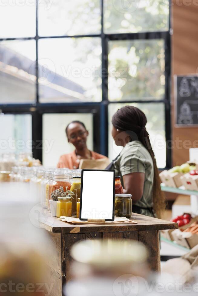 White screen on tablet displays an ad template promoting fresh, organic and sustainable food products in an eco-friendly marketplace. Vendor speaks with client while device shows blank mockup. photo