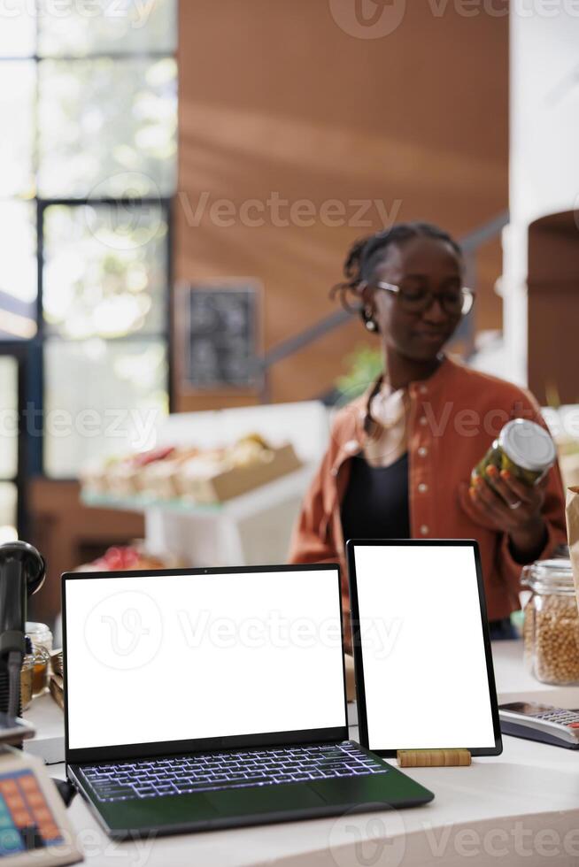 Client looking at pickle jar at counter, with a laptop and tablet displaying empty white screens. Digital devices present a blank chromakey template suitable for promoting an eco friendly bio store. photo