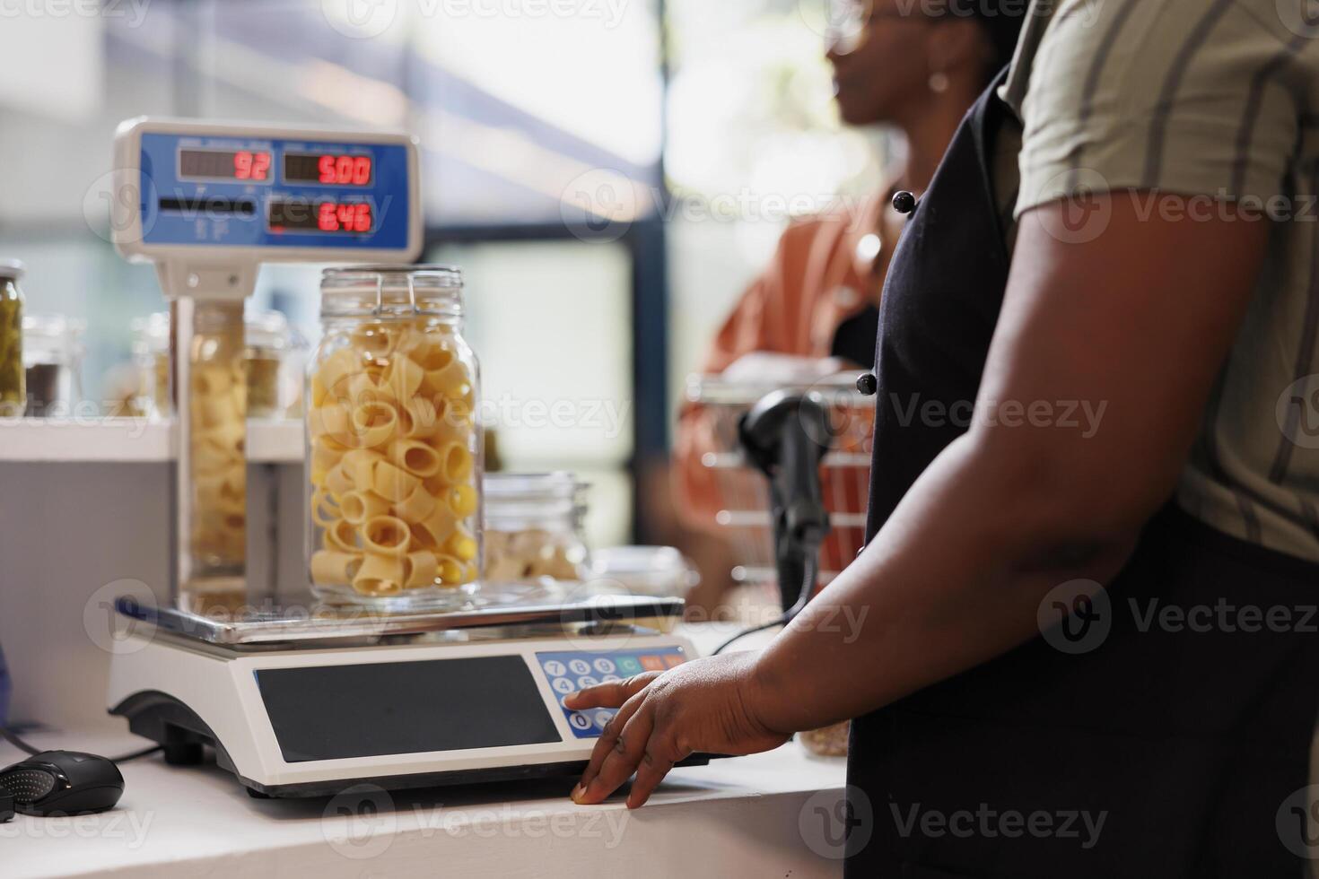 Detailed shot of seller utilizing digital scale to determine weight of container filled with organic produce. Closeup of vendor weighing jar of fresh pasta, with client patiently waiting at counter. photo