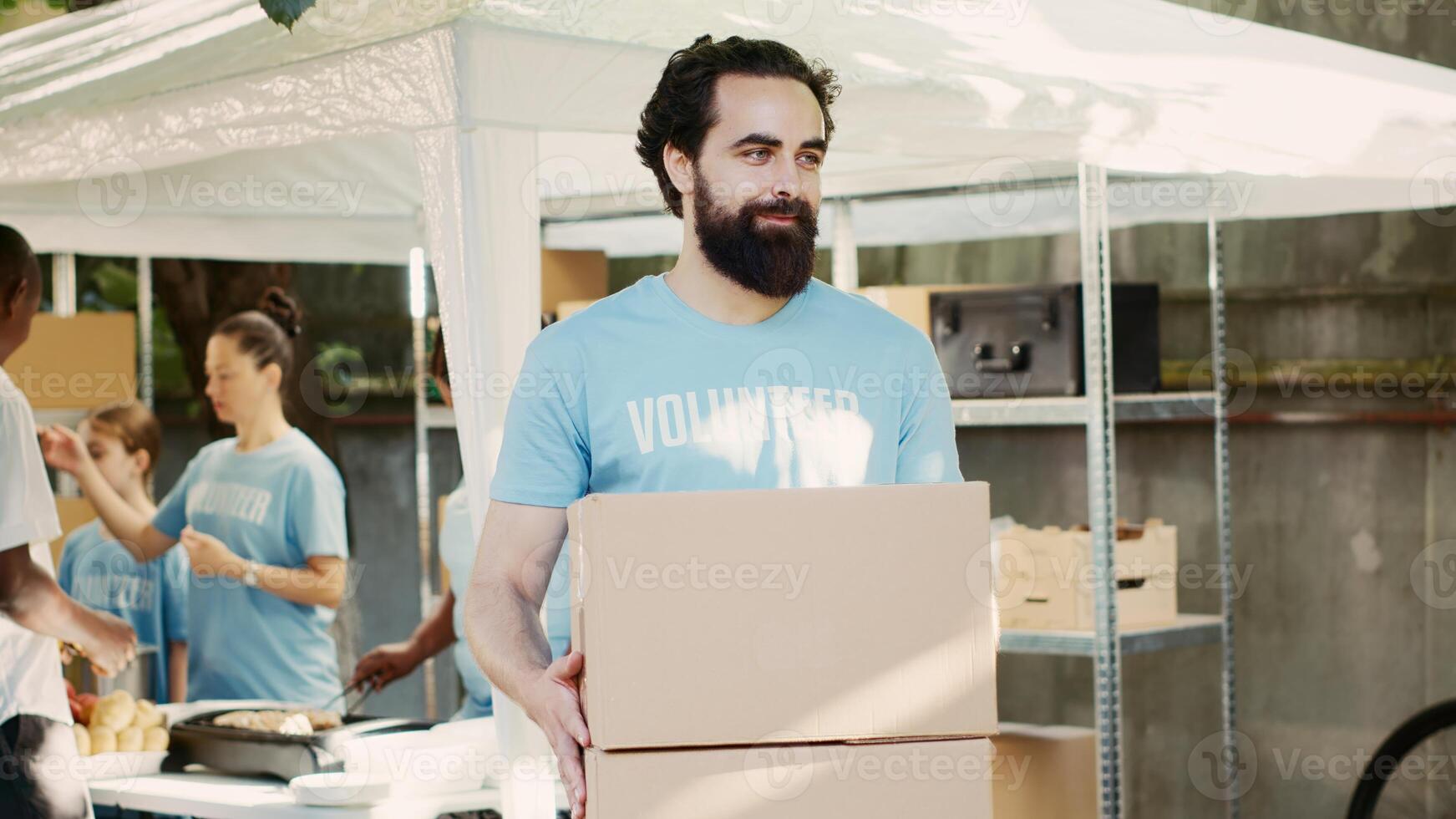 Kind young man donates non-perishable food items to the hungry and homeless as part of community food drive. Male caucasian volunteer carrying contribution boxes while looking at camera. Tripod shot. photo