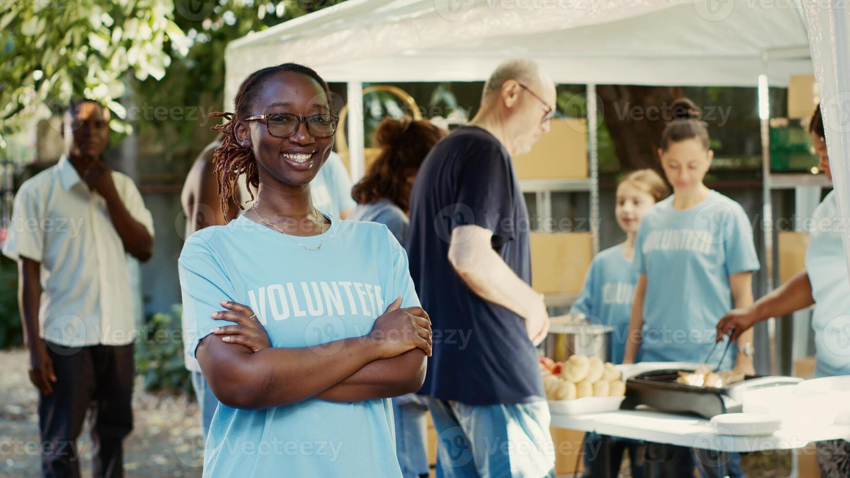 Black lady with spectacles having her arms crossed and gaze fixed on the camera outdoors. Charity workers contributing to non-profit program committed to assisting homeless people. Tripod portrait. photo