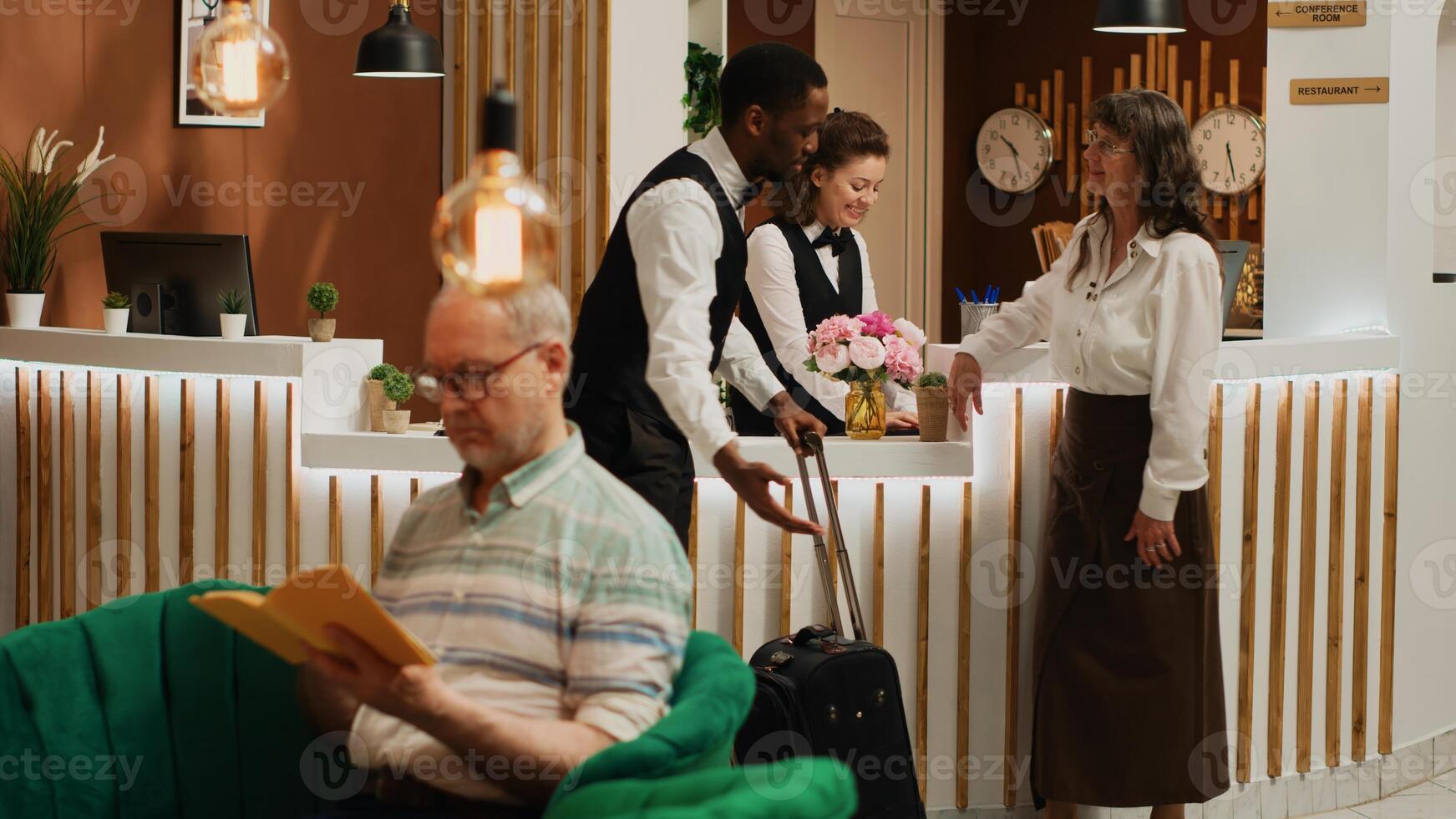 Older guest arriving on vacation at hotel with room reservation, bellhop offering concierge services carrying baggage for tourist. Receptionist doing check in with woman at front desk. Tripod shot. photo
