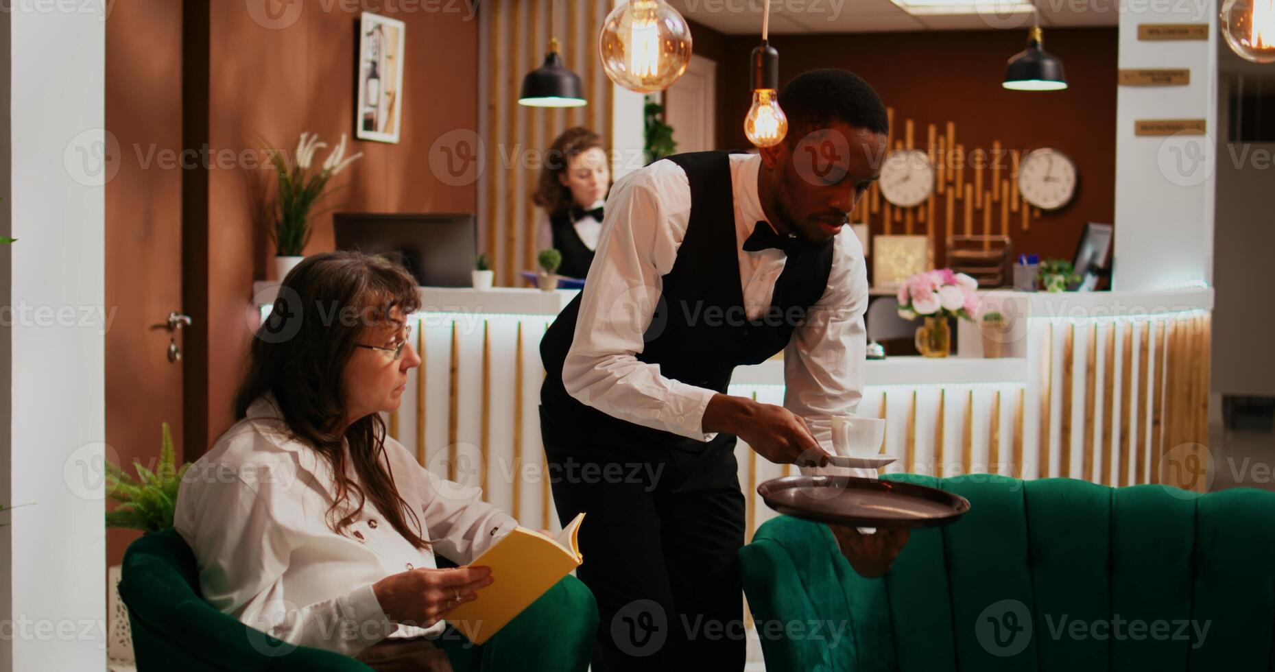 Bellhop serving coffee cup to woman client relaxing in lounge area, senior hotel guest enjoying refreshment and reading book. Employee providing all inclusive services on holiday. photo