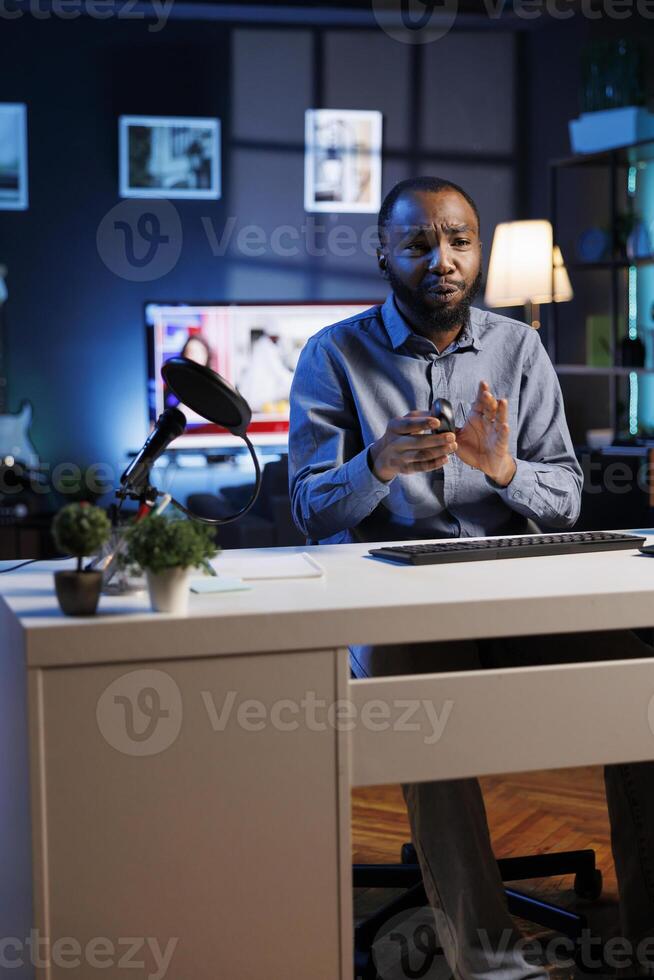 African american man filming video for online channel in dimly lit home studio, presenting gaming mouse to audience. Content creator showing viewers computer peripheral device photo
