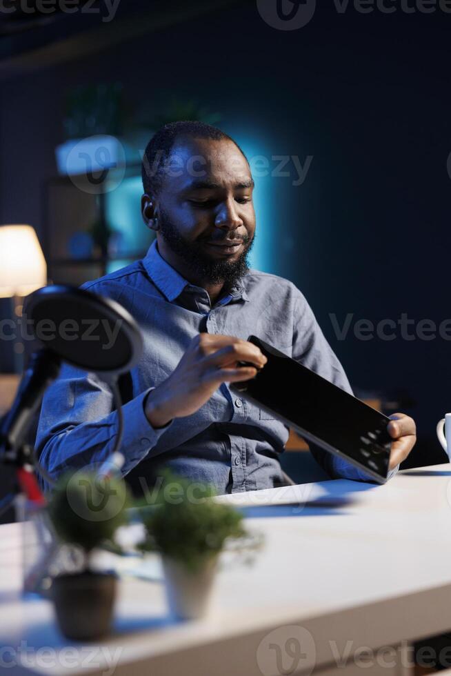 Tech engineer presenting tablet in entry level low budget price range, filming from dimly lit home studio. Viral tech content creator reviewing digital device in front of online audience photo