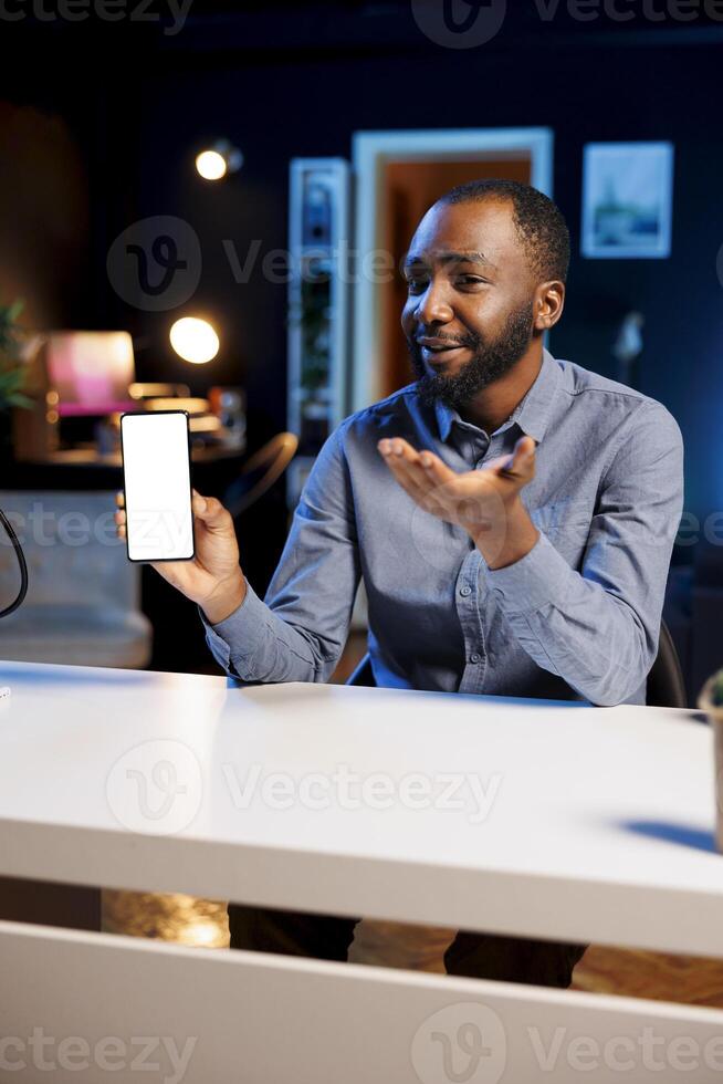 BIPOC content creator holding isolated screen smartphone in his hands, talking about specifications to online audience. Internet show host presenting mockup mobile phone features photo