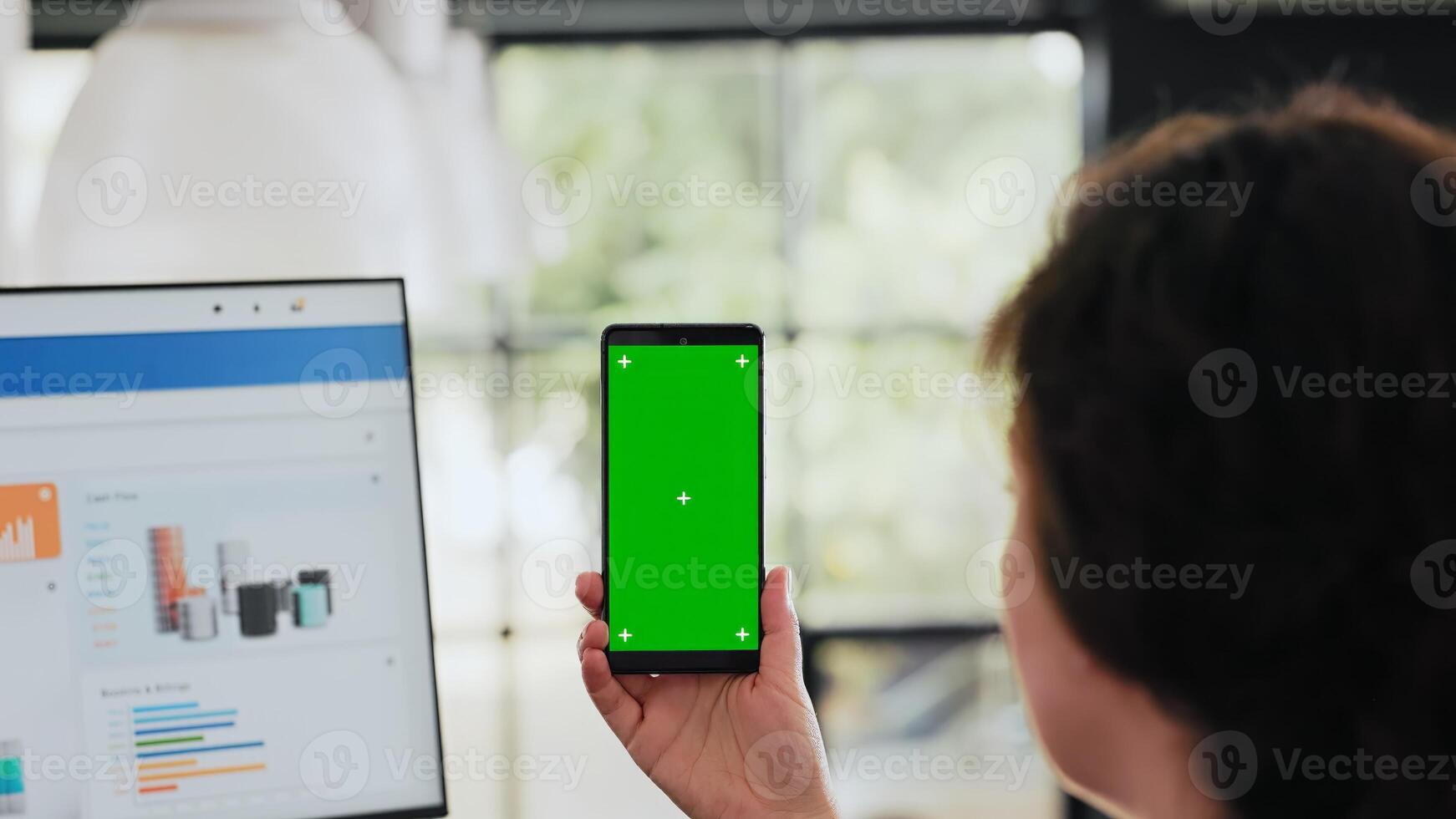 Worker examines phone with greenscreen sitting at workstation in business agency office, analyzing blank chromakey display with copyspace layout. Person works on smartphone. Handheld shot. Close up. photo