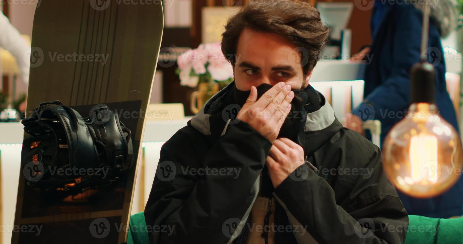 Athletic man getting ready to ski on alpine slopes, sitting in lobby at winter resort before taking snowboard for extreme activity. Hotel guest dressed in skiing suit with protective headgear. photo
