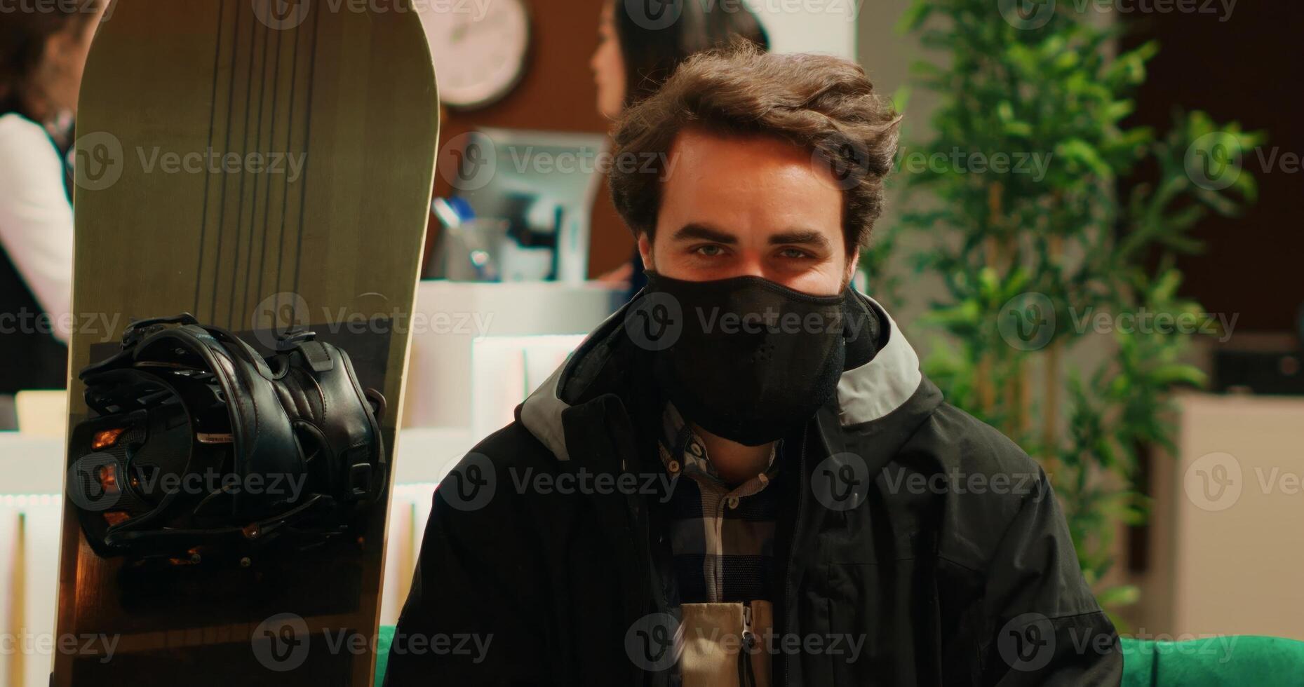 Active person equipped for ski on snowy slopes, sitting in winter complex lobby before taking snowboard on mountain. Professional snowboarder with scarf, protective helmet and outerwear. photo