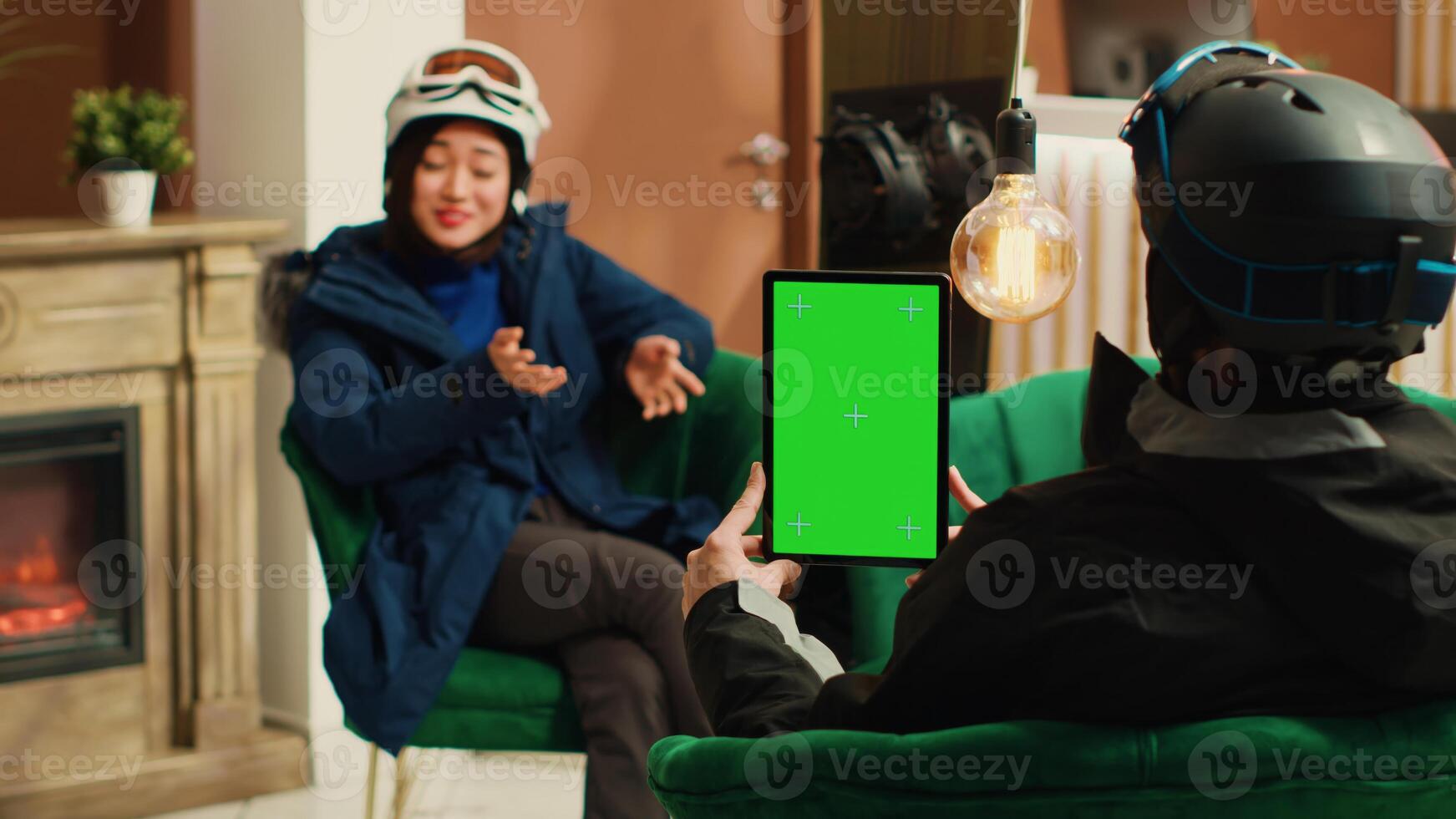 Traveller shows isolated greenscreen on tablet while she sits with skiing partner in lounge area, preparing for winter sport with gear. Man holding gadget with blank chromakey mockup template. photo